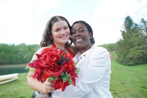 Two women embrace joyfully outdoors, with one holding a bouquet of red roses. The background features greenery and a body of water, suggesting a celebratory occasion.