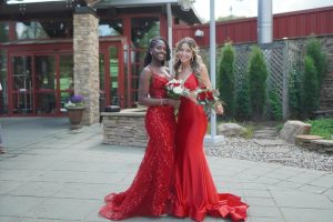 Two women in elegant red dresses pose together outdoors, smiling and holding a bouquet. The background features a rustic building and greenery.