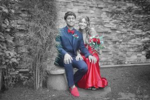 A young couple in red and blue formal attire sits joyfully on a stone bench against a brick wall, surrounded by lush greenery. The woman holds a vibrant bouquet.