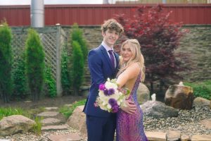 A young couple poses together in a garden setting, dressed elegantly for a special occasion. The woman holds a bouquet of flowers, and they both smile happily.