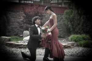 A man kneels with a bouquet, presenting it to a woman in an elegant red dress, surrounded by a garden setting. The scene conveys a romantic moment.