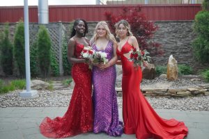 Three young women pose together outdoors, each wearing elegant evening gowns in vibrant colors, holding bouquets of flowers. The setting features greenery and decorative stones in the background.