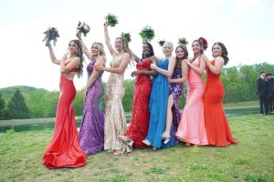 A group of eight women in colorful evening gowns poses together outdoors, each holding a bouquet. They display joyful expressions, celebrating a special occasion.