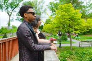 A young man in a stylish black suit and sunglasses stands beside a woman in a formal dress, both gazing thoughtfully into the distance. They are surrounded by lush greenery in a park setting.