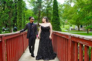A couple walks hand in hand across a wooden bridge surrounded by lush greenery, with the woman in an elegant black gown and the man in a formal suit. The scene captures a moment of connection and celebration.