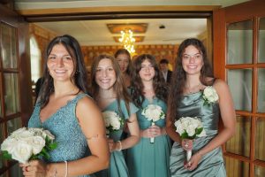 Four young women in teal dresses stand together, each holding a bouquet of white roses, smiling as they pose in a warmly lit room. The atmosphere suggests a celebratory occasion, likely a wedding or formal event.