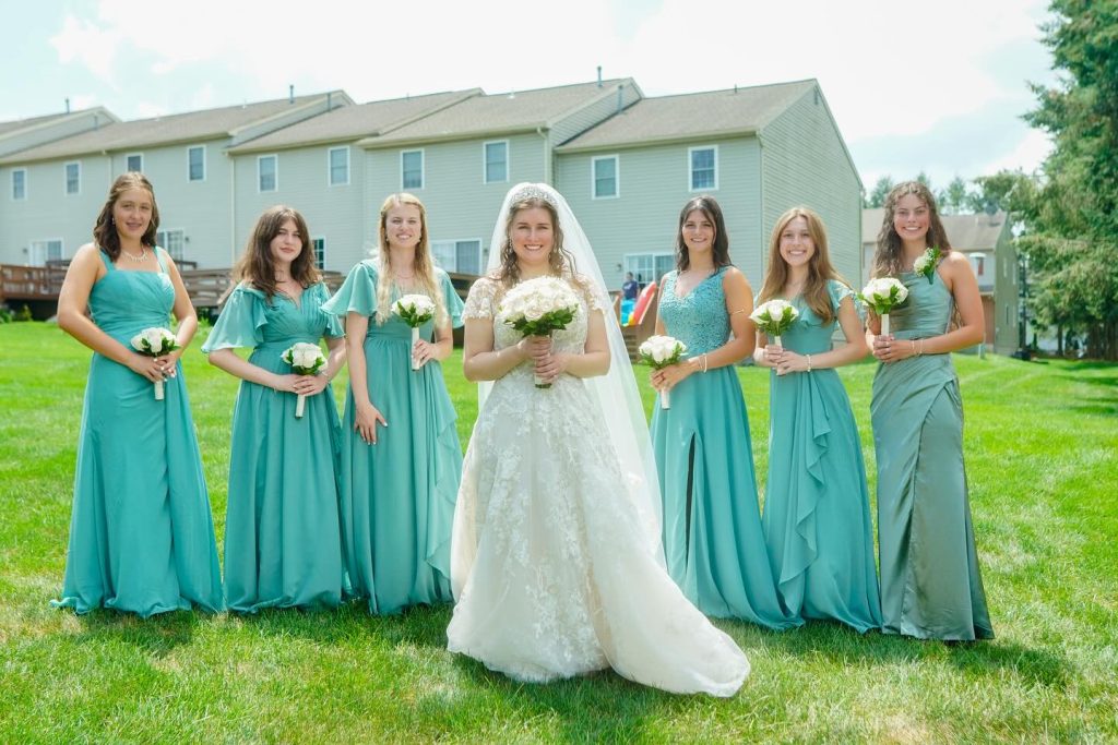 A bride stands in the center, surrounded by her bridesmaids, all wearing teal dresses and holding bouquets. The setting is a grassy area with houses in the background.