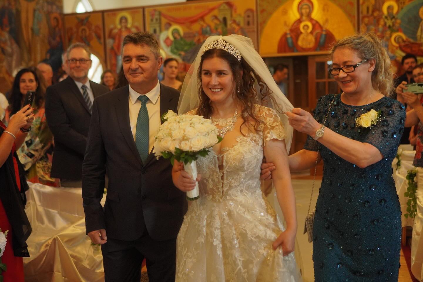 A bride in a beautiful white gown holds a bouquet of flowers while being escorted by her father and another woman in a decorated venue. The background features colorful religious icons, suggesting a wedding ceremony.
