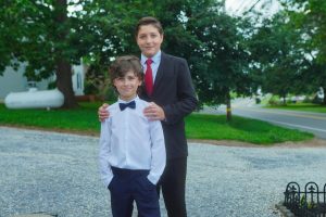 Two boys dressed in formal attire stand together outdoors, with one boy in a white shirt and bow tie and the other in a suit and tie. The background features greenery and a gravel path.