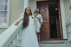 A young girl in a white dress smiles on a staircase outdoors. Two other children stand behind her at a wooden door. The scene is joyful and bright.