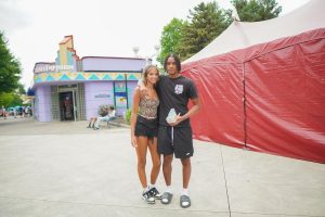 A young couple poses together in a colorful amusement park setting, with a vibrant building and a large tent in the background. They both appear cheerful and relaxed.