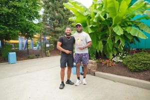 Two men stand together in a lush outdoor setting, surrounded by green plants and colorful flowers. They are smiling and dressed casually, enjoying a sunny day.