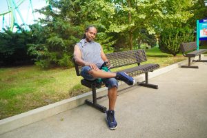 A man is sitting on a park bench, casually checking his phone while surrounded by greenery. The scene conveys a relaxed atmosphere.
