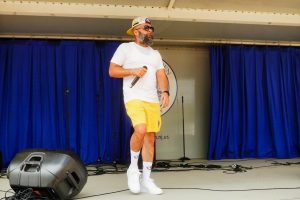 A man in a white t-shirt and yellow shorts performs on stage, holding a microphone, with a blue curtain in the background. The scene captures the energy of a live performance.