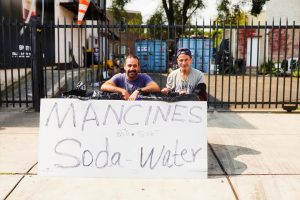 Two men sit behind a large sign that reads "MANCINES Soda Water," with a fence and some containers visible in the background. The scene appears to be a casual outdoor setup, likely for selling beverages.