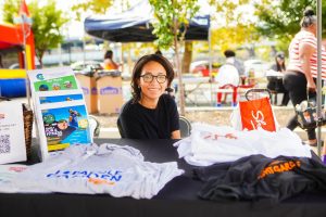 A young woman smiles while sitting behind a table displaying various T-shirts at an outdoor event. Colorful tents and people can be seen in the background, creating a lively atmosphere.
