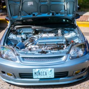 A silver car with its hood open, showcasing a polished engine and various modifications. The license plate reads "MONICA."