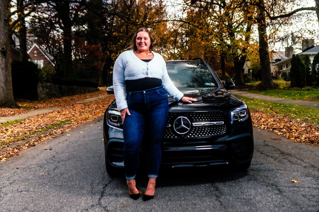 A woman stands confidently next to a black Mercedes-Benz SUV on a tree-lined street covered in autumn leaves. The scene captures a vibrant fall atmosphere.