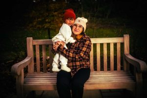 A woman sits on a wooden bench holding a young child, both wearing warm hats, in a natural outdoor setting. The scene conveys a sense of warmth and affection.