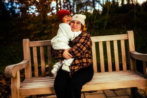 A woman sits on a bench in a park, holding a child who is smiling and wearing a red hat. The scene is set against a backdrop of autumn foliage.