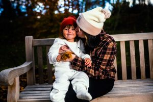 A mother kisses her child on the cheek while sitting on a bench in a park, surrounded by autumn foliage. The child wears a red hat and a white outfit, and they both share a joyful moment together.
