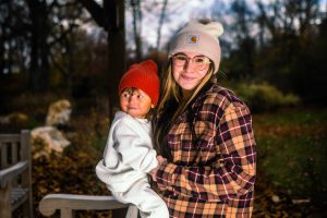 A young woman holds a smiling child in a cozy outdoor setting, both wearing warm hats. The background features trees and autumn foliage.