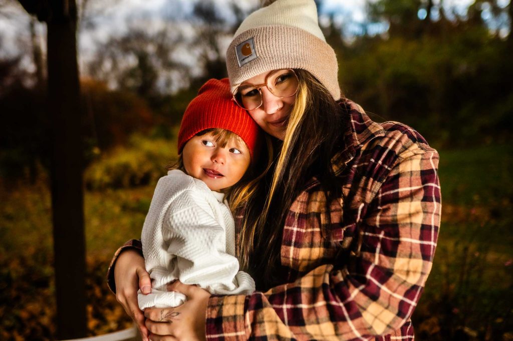 A woman with long hair and glasses smiles while holding a young child wearing a red beanie. They are surrounded by a natural setting with autumn colors.