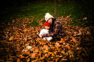 A woman and a child sit together in a pile of autumn leaves, both wearing warm hats. The scene captures a cozy moment in a colorful fall setting.