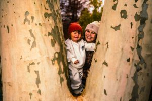 A woman and a child are playfully peeking out from between the trunks of two trees, both smiling and wearing warm hats. The scene captures a joyful moment in a natural setting.