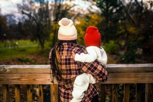 A woman wearing a plaid coat and a beanie holds a child in a red hat, both looking out over a scenic view of trees and greenery. The image captures a moment of connection between them in a natural setting.