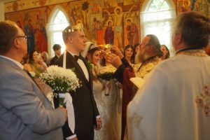 A couple is being crowned during a wedding ceremony, surrounded by guests and a priest in a decorated church. The scene captures a moment of celebration and tradition.