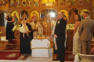 A wedding ceremony taking place in a church, with the couple standing at the altar surrounded by guests and a priest. The setting features ornate religious icons in the background.