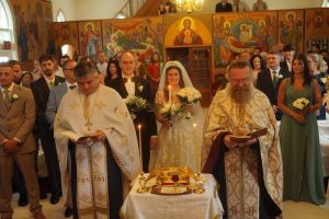 A bride and groom stand at the altar during a wedding ceremony, surrounded by clergy and guests in a decorated church. The atmosphere is solemn and celebratory, with candles lit and floral arrangements present.