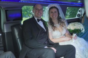 A couple dressed for a wedding sits together in a limousine, with the bride holding a bouquet of flowers and wearing an elegant gown. The interior is illuminated with soft blue lighting.