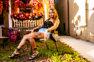 A young woman with tattoos sits confidently on a chair in a garden, surrounded by vibrant flowers and greenery. The warm sunlight highlights her relaxed pose and stylish outfit.