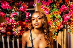 A young woman with long, wavy hair stands in front of a vibrant backdrop of colorful flowers, exuding confidence. The setting is bright and lively, enhancing the overall atmosphere.