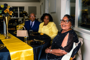 A group of three people smiles while seated at a elegantly decorated table during a celebration, featuring black and yellow decor. The atmosphere suggests a joyful gathering.