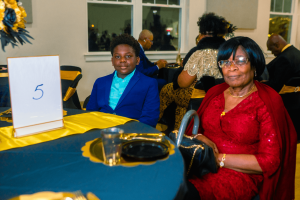 A young boy in a blue suit sits next to a woman in a red dress at a decorated table with a number sign. The setting appears to be a formal event or celebration.