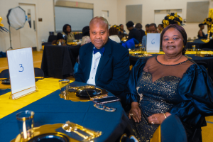 A couple dressed elegantly sits at a formal dining table, surrounded by other guests in a festive atmosphere. The table is decorated with gold and blue accents.