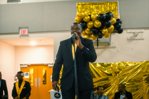 A man in a suit speaks into a microphone at a celebratory event, with a backdrop of gold and black decorations. Guests are seated in the foreground, engaged in the occasion.