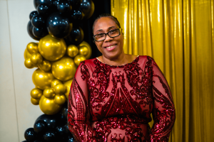 A woman in a red dress with intricate detailing smiles in front of a backdrop of gold and black balloons. The atmosphere suggests a festive or celebratory event.