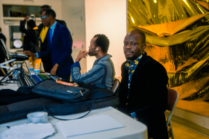 A group of men work at a table with equipment and laptops. One man faces the camera, wearing a patterned shirt. Background features golden decor.
