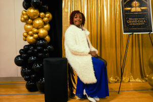 A woman with curly hair is seated in front of a golden backdrop, wearing a white fur coat and blue pants, next to a black and gold balloon decoration. A sign for a gala is visible in the background.