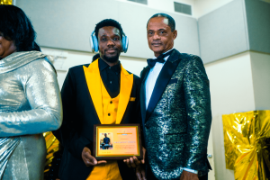 A man wearing a yellow and black outfit and headphones stands proudly holding an award, next to another man in a shiny silver suit. The setting appears to be a celebratory event.