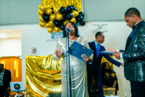 A woman in a silver gown speaks into a microphone at a festive event, while two men stand nearby, one holding a document. The background features gold and black balloons and a shiny gold backdrop.