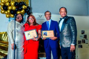 Four individuals stand together at a celebratory event, each holding a certificate. The backdrop features gold and black balloons, adding to the festive atmosphere.