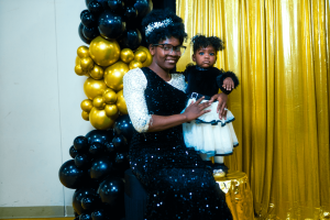 A person in a sparkling black and white outfit holds a small child dressed in a white dress, standing in front of a backdrop of black and gold balloons. The setting appears festive and celebratory.