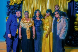 A group of six people dressed in formal attire poses together in front of a colorful backdrop adorned with balloons. The setting appears to be a celebration or event.