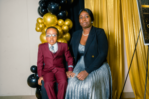A woman in formal attire sits next to a young boy dressed in a suit, both smiling in front of a backdrop of gold and black balloons. --- A woman in formal attire sits next to a young boy dressed in a suit, both smiling in front of a backdrop of gold and black balloons.