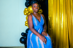 A young woman sits gracefully in a blue dress, surrounded by a festive backdrop of black and gold balloons. The setting suggests a celebration or special event.